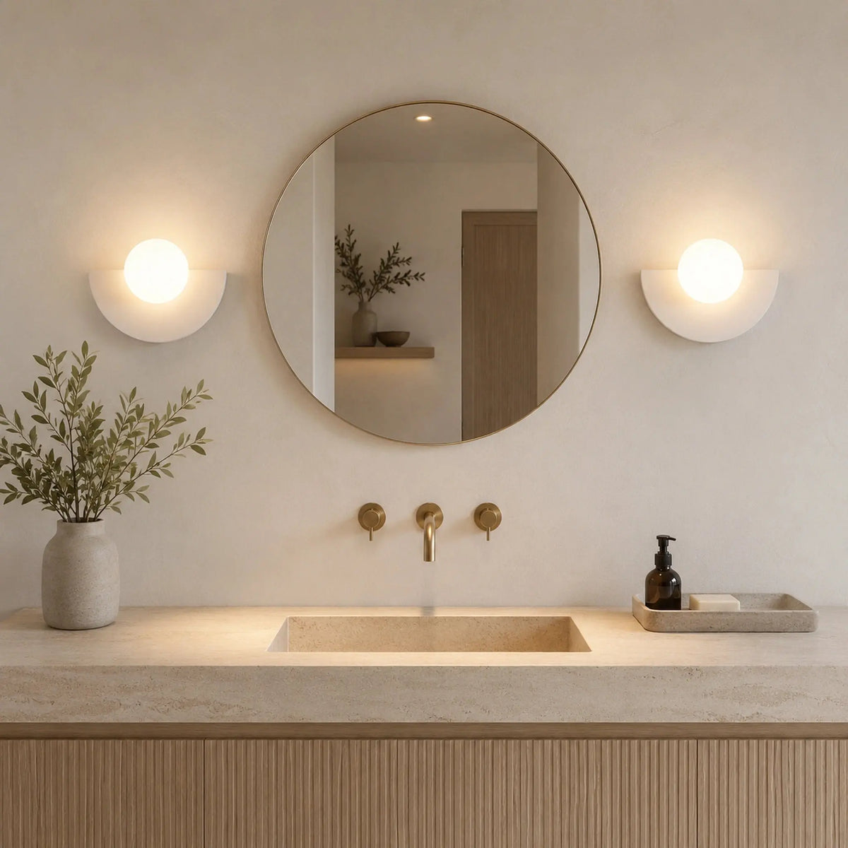 Deux abat-jour muraux blancs en demi-lune encadrant un miroir rond doré dans une salle de bain épurée